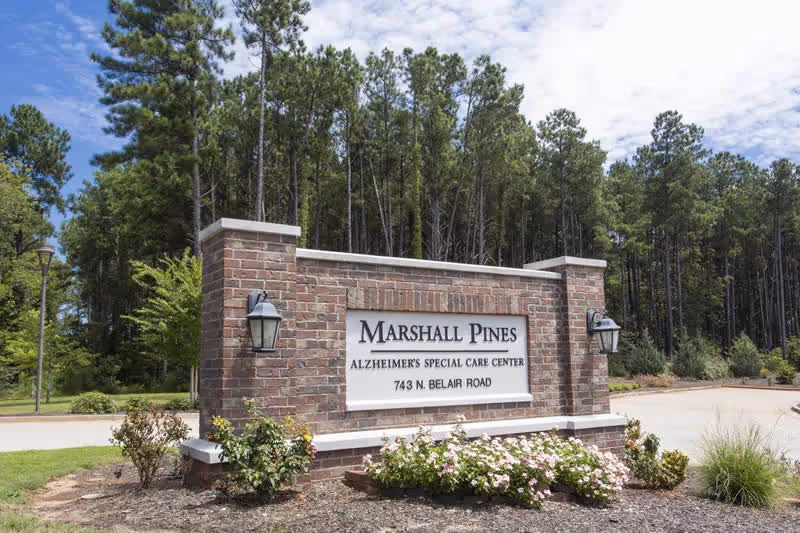 Brick entrance sign for Marshall Pines Alzheimer's Special Care Center (743 N. Belair Road) with landscaping and tall pine trees behind it.