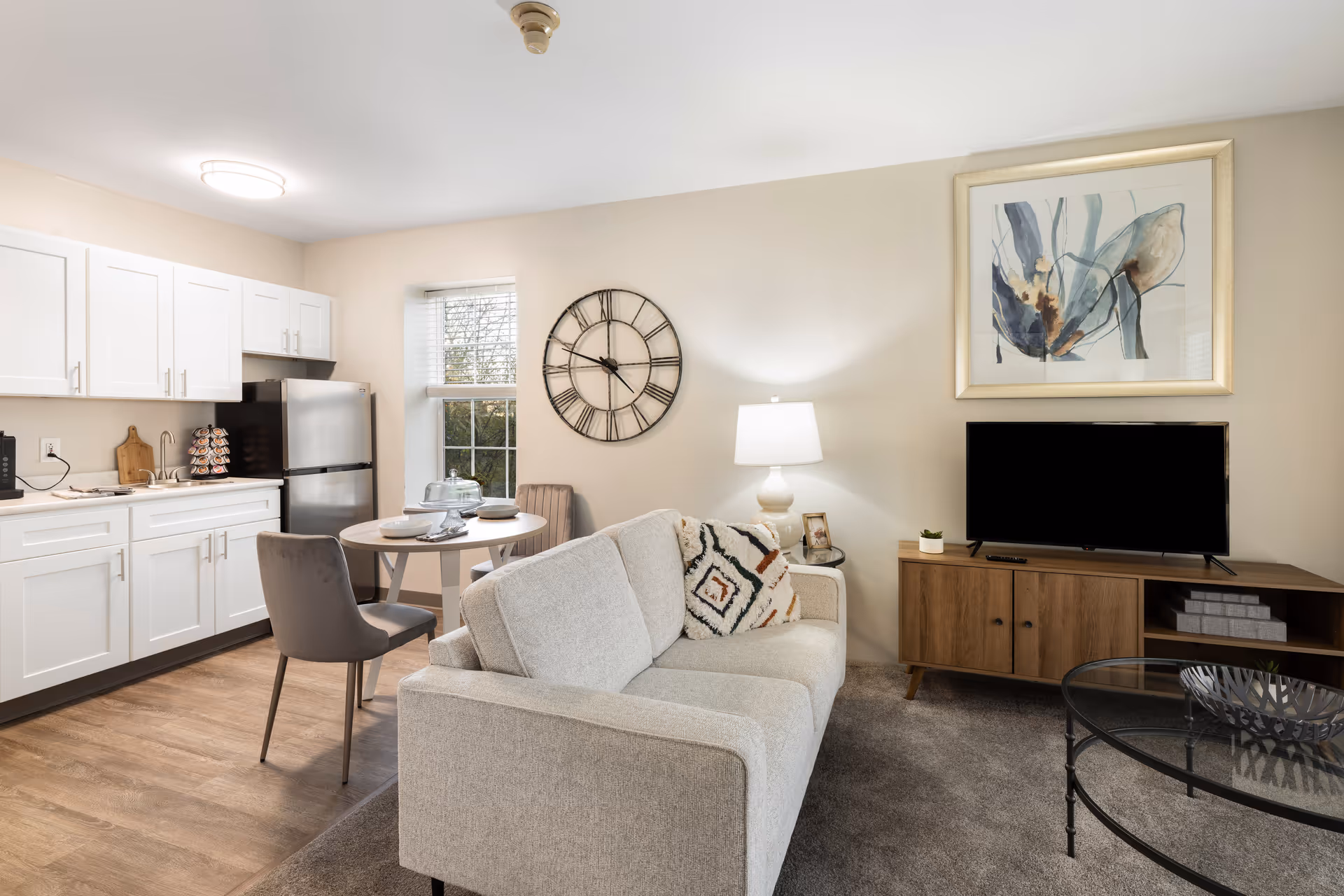 A cozy and modern living area in a senior living community featuring a light gray sofa with patterned cushions, a wooden TV stand with a flat-screen television, a glass coffee table, a round dining table with two gray chairs, and a kitchen area with white cabinets, a stainless steel refrigerator, and a window letting in natural light. The walls are decorated with a large clock and an abstract framed painting.
