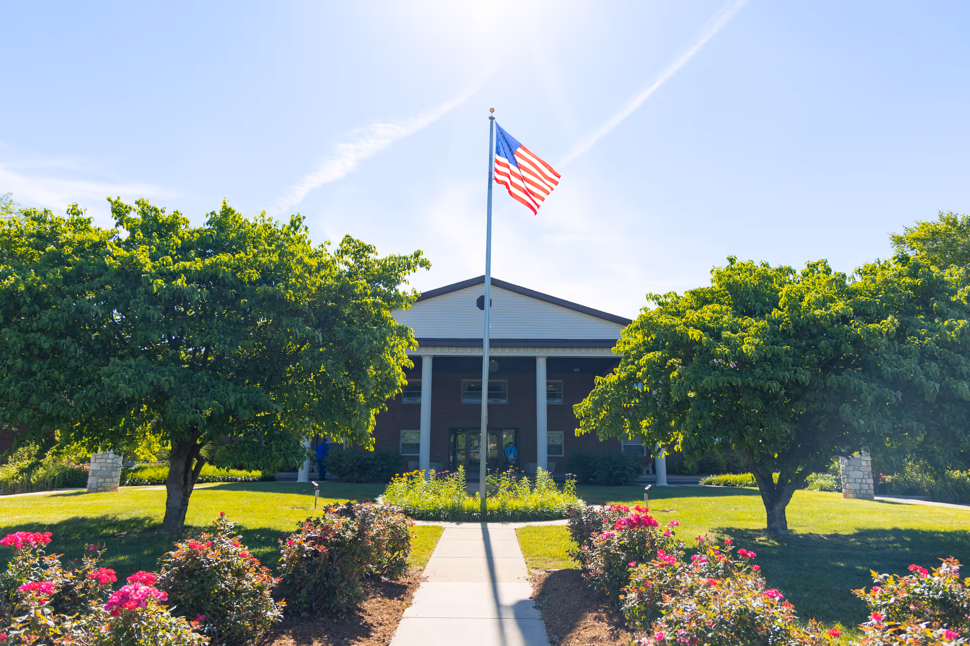 Front entrance of a senior living building with an American flag on a pole, flanked by trees and flowerbeds.