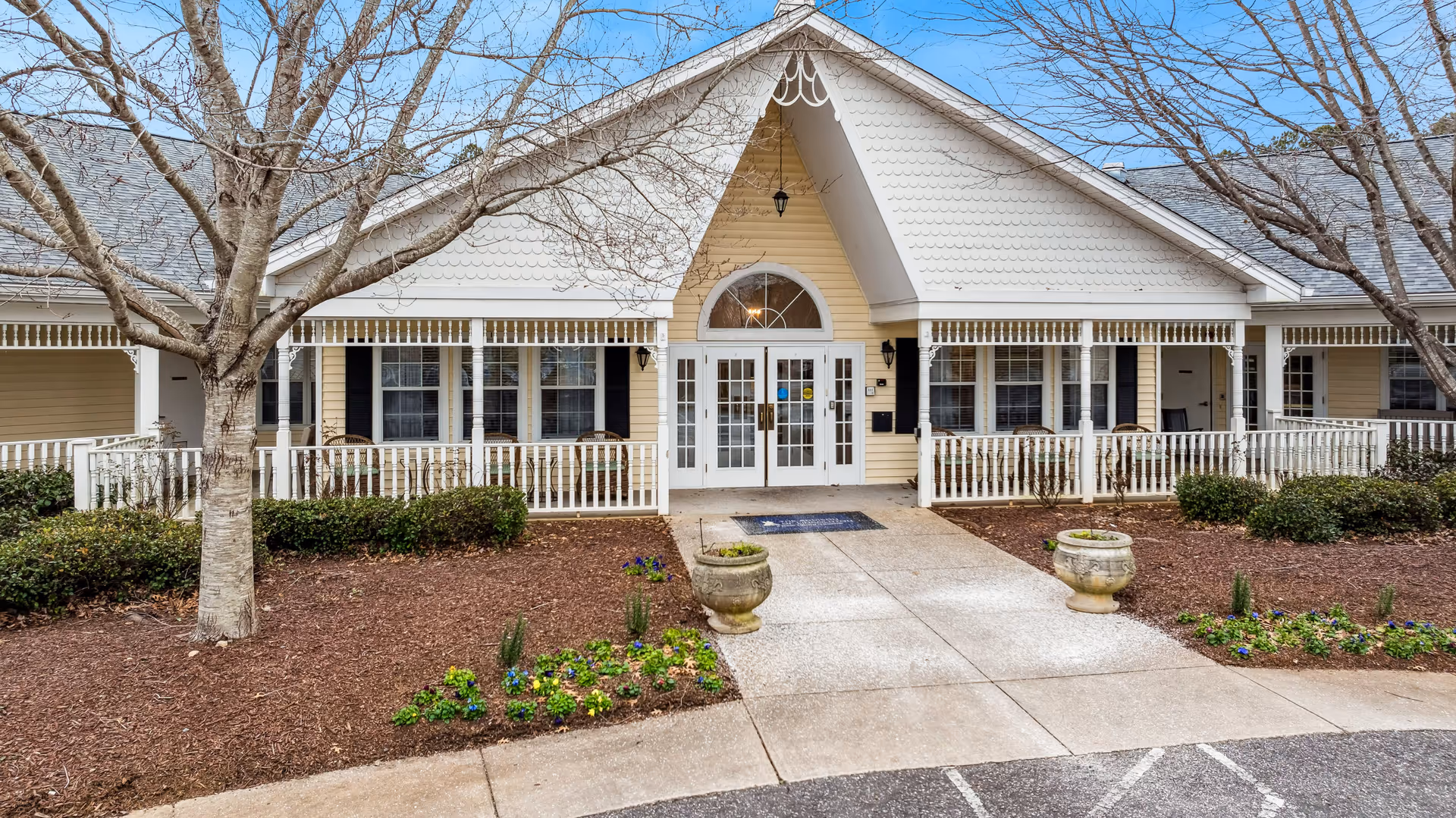 Front exterior view of a single-story building with a peaked roof and a covered porch. The building is painted yellow with white trim and has multiple windows with black shutters. There are two large planters with flowers near the entrance, a concrete walkway leading to double glass doors, and leafless trees and shrubs in the landscaped area in front.