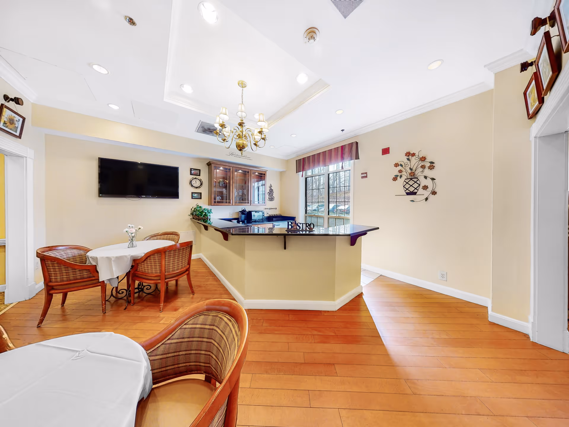 A bright dining area in a senior living facility with wooden floors, a small round table covered with a white tablecloth and surrounded by four wicker chairs. There is a counter labeled 'Bistro' with a coffee machine and a glass cabinet behind it. A flat-screen TV is mounted on the wall, and a window with a valance lets in natural light. The walls are decorated with framed pictures and a metal wall art piece shaped like a vase with flowers.