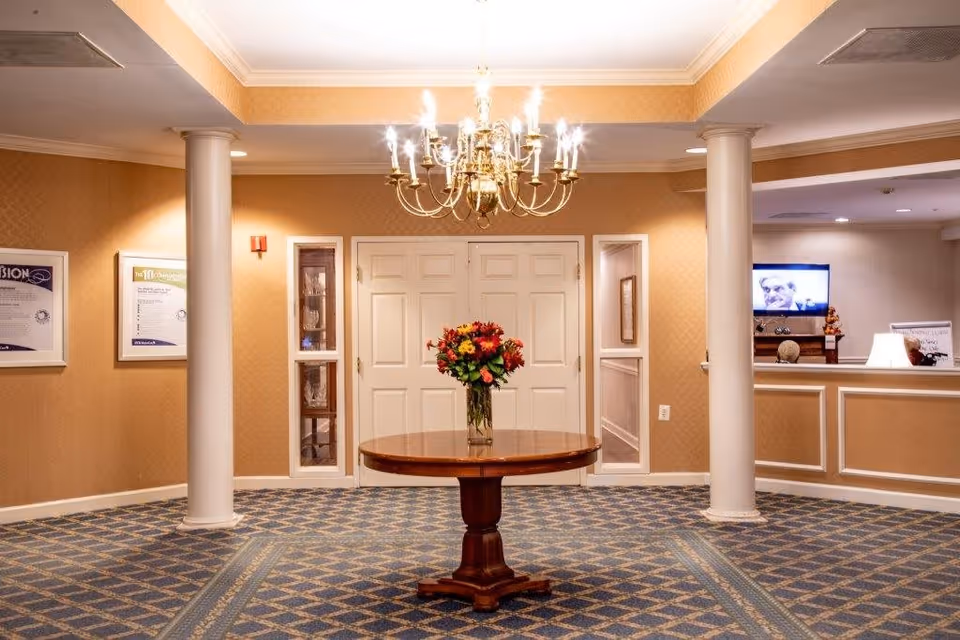 Interior view of a senior living facility lobby area with a round wooden table in the center holding a vase of colorful flowers. The room has patterned carpet flooring, beige walls, white columns, a chandelier hanging from the ceiling, and a reception desk with a television in the background.