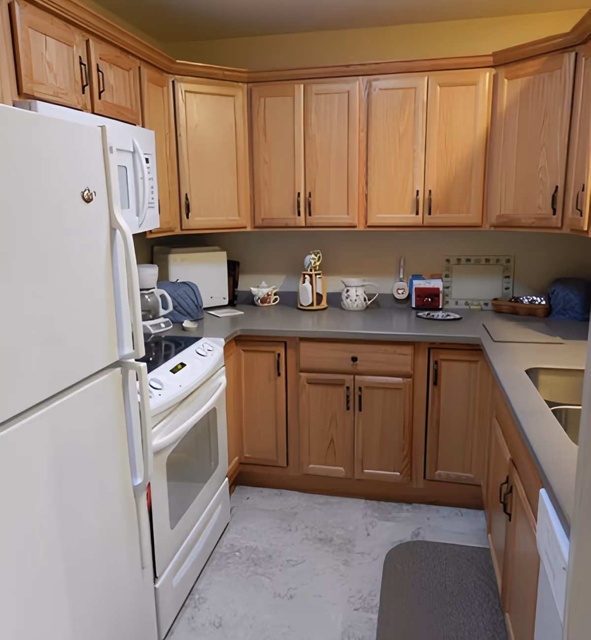 A compact kitchen with light wood cabinets, a white refrigerator, white stove with oven, microwave, coffee maker, and various kitchen items on the gray countertop.
