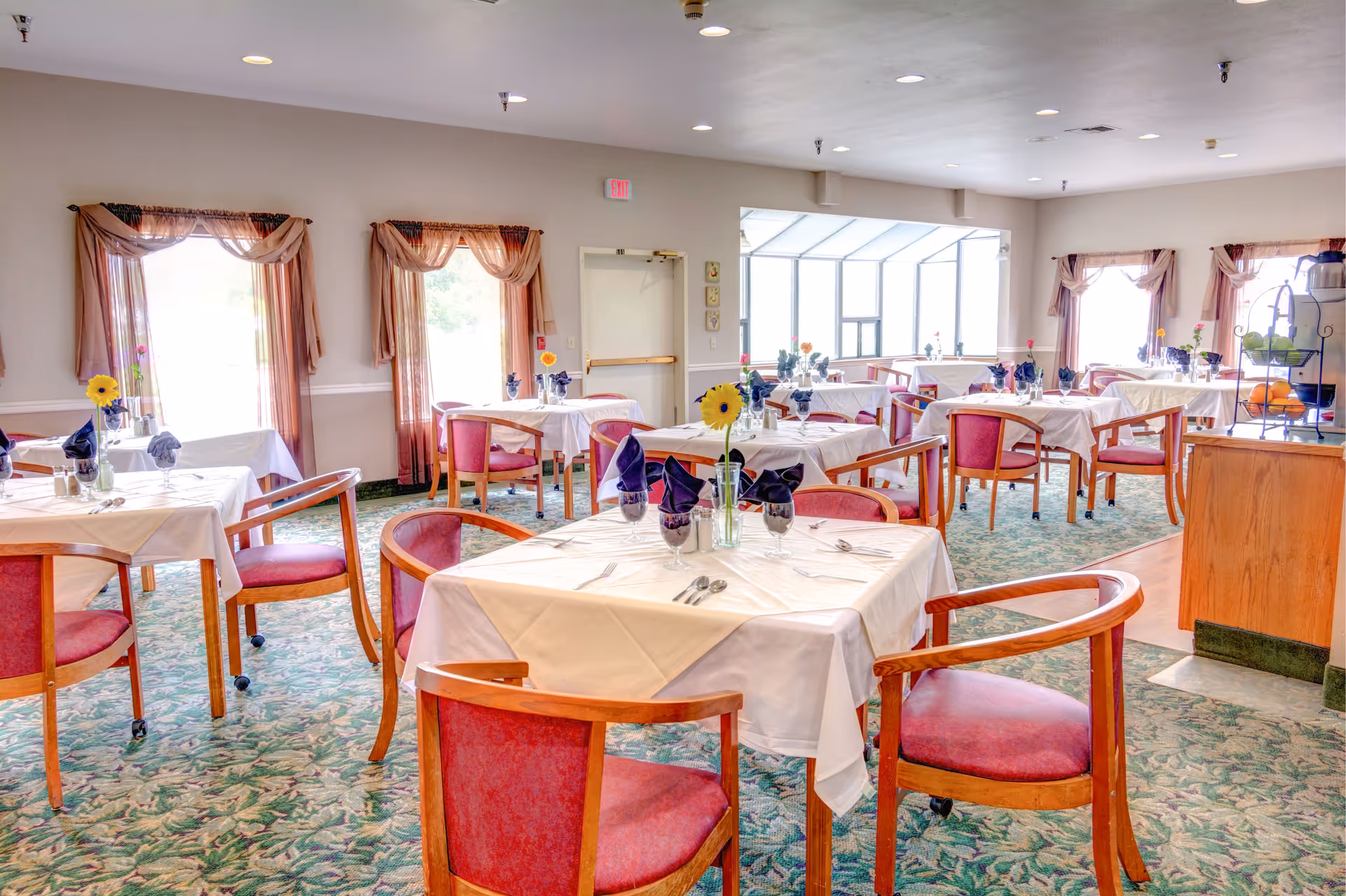 Bright dining room with multiple tables set with white linens, napkins, flowers, and wooden chairs.