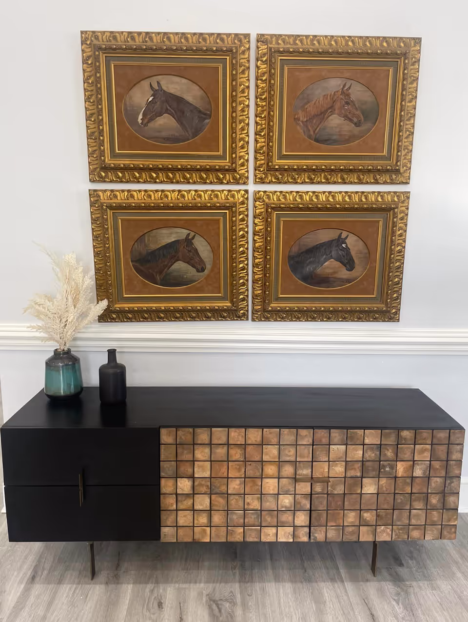 A modern black and wood-textured sideboard with two decorative vases on top, placed against a white wall. Above the sideboard are four framed paintings of horse heads, each in an ornate gold frame arranged in a 2x2 grid.