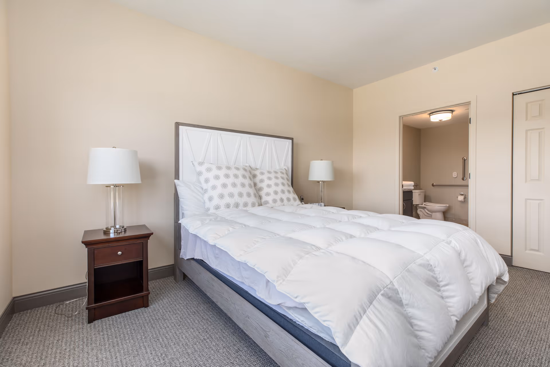 A bright, furnished bedroom with a white comforter, patterned pillows, nightstands and lamps, and an open doorway to a bathroom.