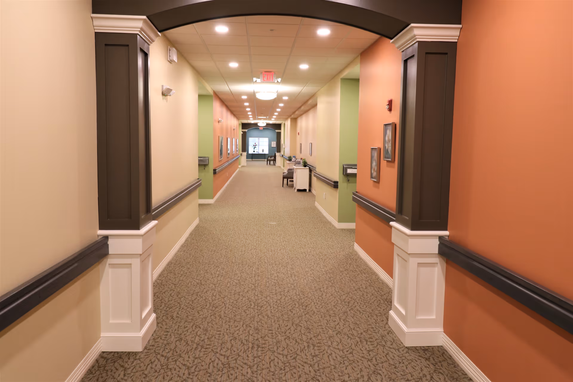 A long, well-lit hallway in a senior living facility with beige, orange, and green walls, carpeted floor, handrails along both sides, decorative columns, framed pictures on the walls, and chairs and small tables placed along the corridor.