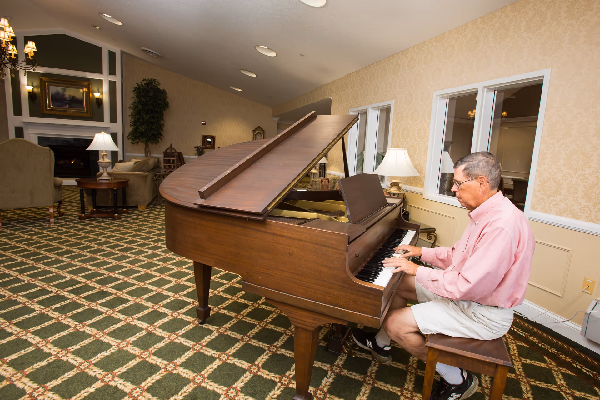An elderly man wearing a pink shirt and white shorts is playing a grand piano in a spacious, well-lit living room with patterned carpet, beige wallpaper, and comfortable seating including armchairs and a sofa. There are lamps on side tables and a fireplace with a framed painting above it in the background.
