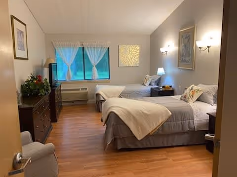 A cozy bedroom with two single beds, each made with gray bedding and white blankets. There is a nightstand with a lamp between the beds, wall-mounted lights above, and framed artwork on the walls. A window with sheer white curtains lets in natural light. A dresser with a TV and a floral arrangement is on the left side, and a cushioned chair is near the door. The room has wooden flooring and a calm, neutral color scheme.