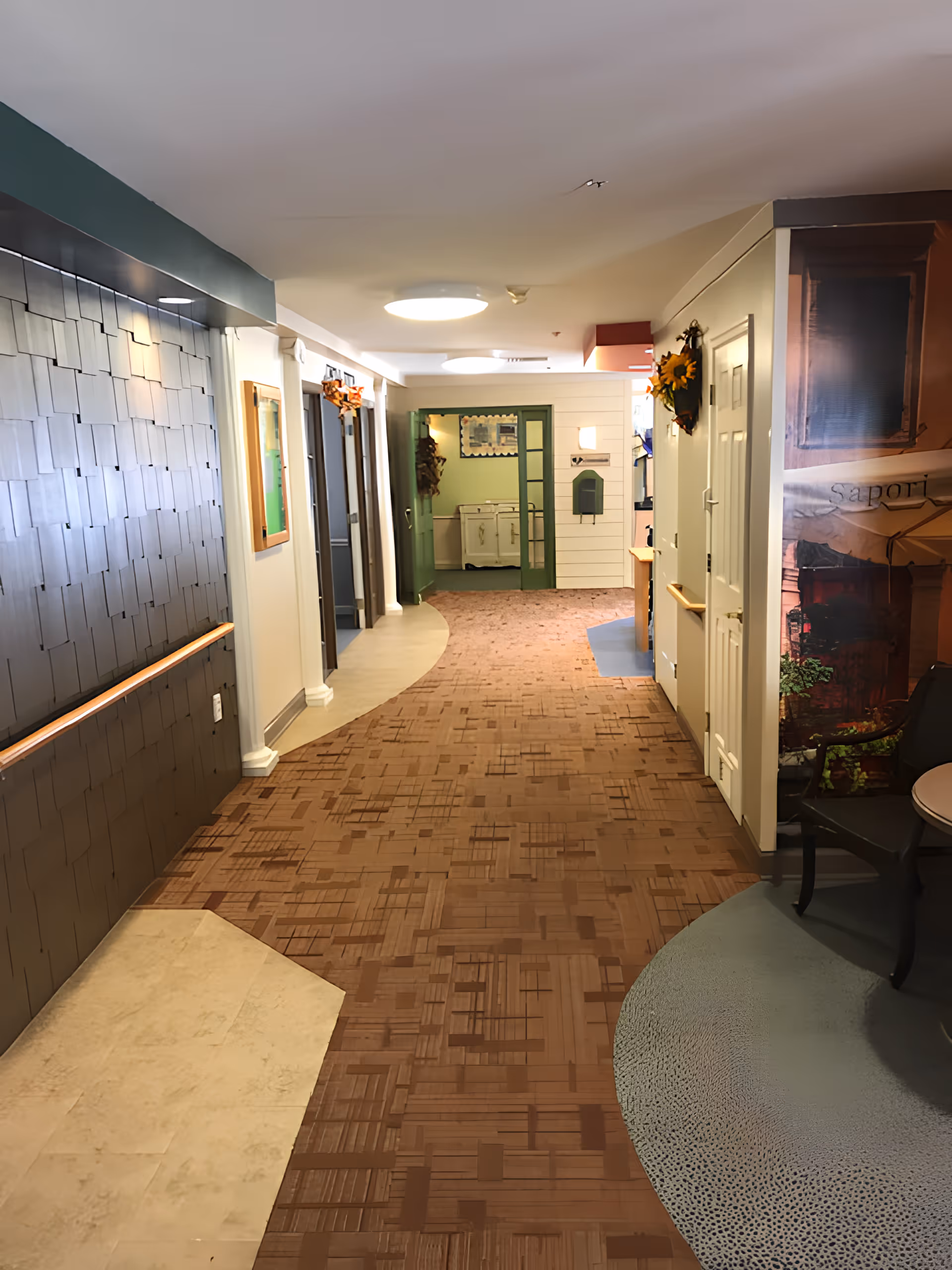 A well-lit hallway in a senior living facility with a patterned carpet floor and light-colored walls. The left wall features a textured panel with a handrail, while the right side has doors and a small seating area with a chair and table. The hallway leads to a room with green double doors and a white cabinet inside. There are decorative wreaths on the walls and ceiling lights providing illumination.