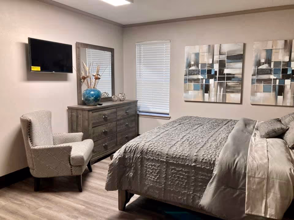 A cozy bedroom with a large bed covered in a textured gray comforter and multiple pillows. There is a gray upholstered armchair in the corner, a wooden dresser with a large mirror above it, and a decorative blue vase with dried plants on top. A flat-screen TV is mounted on the wall, and two abstract paintings hang above the bed. The room has light-colored walls and wood flooring.