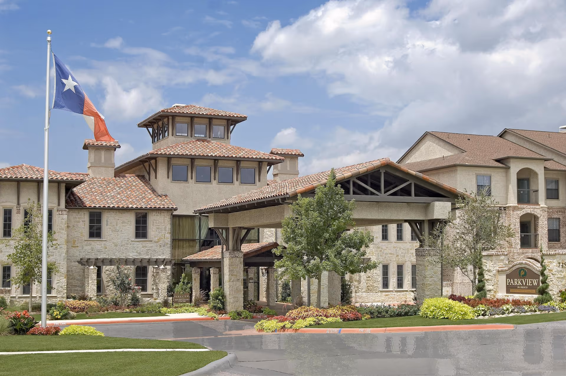Front entrance of the Parkview in Frisco senior living building with a covered porte-cochère, landscaped grounds, and a Texas flag flying nearby.