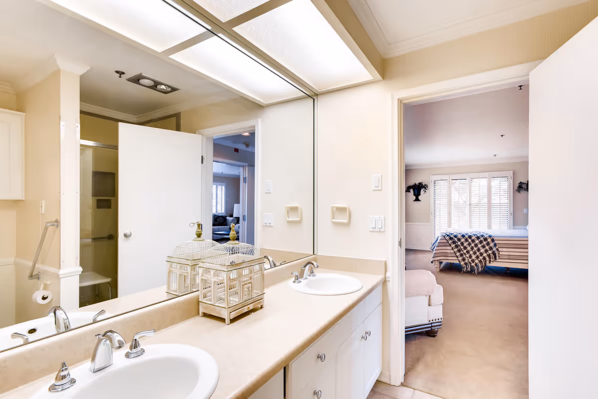 A bright bathroom with a large mirror above a double sink vanity. The countertop is beige with two white sinks and silver faucets. A decorative birdcage is placed on the counter. The bathroom door is open, revealing a bedroom with a bed covered by a striped blanket and a checkered throw, and a window with white blinds. The bathroom also has a shower with a sliding glass door and a grab bar.