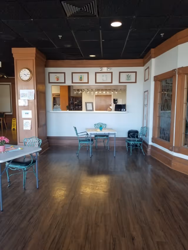 Interior view of a senior living facility common area with wooden flooring and a black ceiling. There are two tables with green metal chairs, one table has a small flower pot with pink flowers. A wooden pillar with a clock and some drawings is visible on the left. The back wall has a serving window with framed botanical prints above it and a kitchen or service area behind the window.
