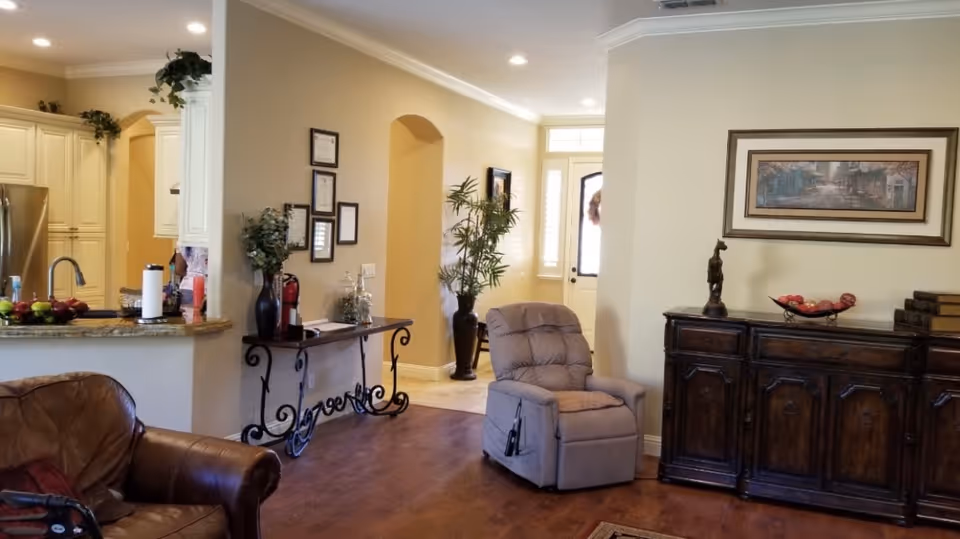 Interior view of a living area in a care home featuring a brown leather sofa, a beige recliner chair, a wooden sideboard with decorative items, a framed painting on the wall, and a kitchen area with cream-colored cabinets and a granite countertop. The space has wooden flooring and neutral-colored walls with some plants and framed certificates or pictures on the wall.