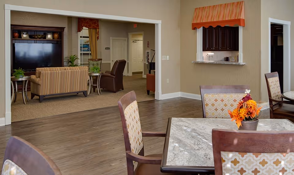 Interior view of a senior living facility showing a dining area with tables and chairs in the foreground, a small kitchen pass-through window with dark cabinets and a striped valance, and a living room area with a large TV, striped sofa, and armchairs in the background.