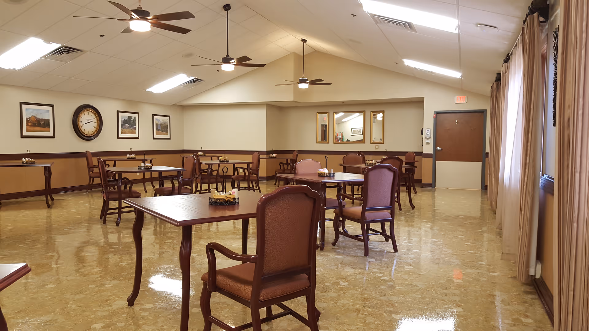 A bright dining room with multiple wooden tables and upholstered chairs, ceiling fans, framed wall art, and large windows with curtains.