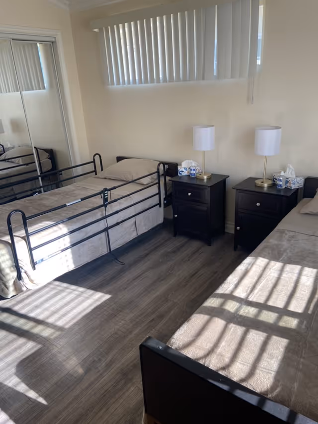 A bedroom with two single beds separated by two dark wooden nightstands, each with a white lamp and tissue boxes. The room has light-colored walls, vertical blinds on the window, and a mirrored closet door reflecting one of the beds. The floor is a dark wood laminate.