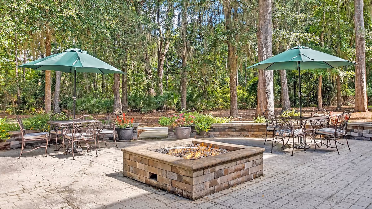 Outdoor patio area with two green umbrellas shading metal tables and chairs, a square brick fire pit in the center, surrounded by trees and greenery.