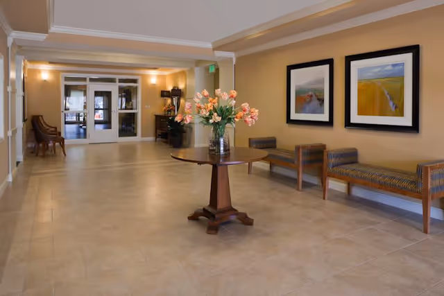 A spacious and well-lit interior hallway with beige tiled floors and light yellow walls. In the center, there is a round wooden table with a vase of pink tulips. On the right side, there are two cushioned benches with patterned upholstery beneath two framed landscape paintings. At the far end, double glass doors lead to another area, with additional seating and a small table with a lamp visible.