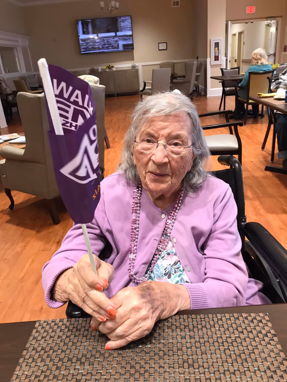 An older woman in a wheelchair holding a small purple flag while seated at a dining table in a senior living common room.