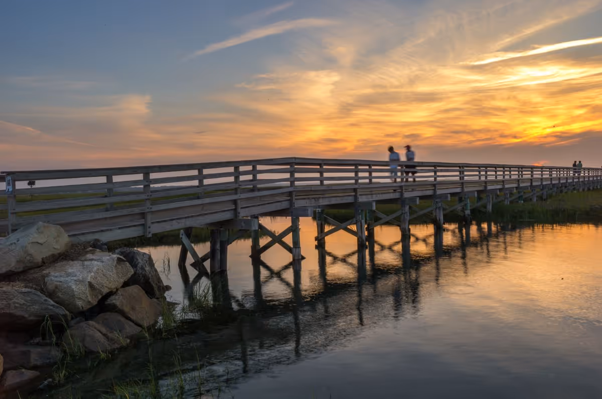 A wooden boardwalk bridge over calm water at sunset with a colorful sky and a few people walking on the bridge.