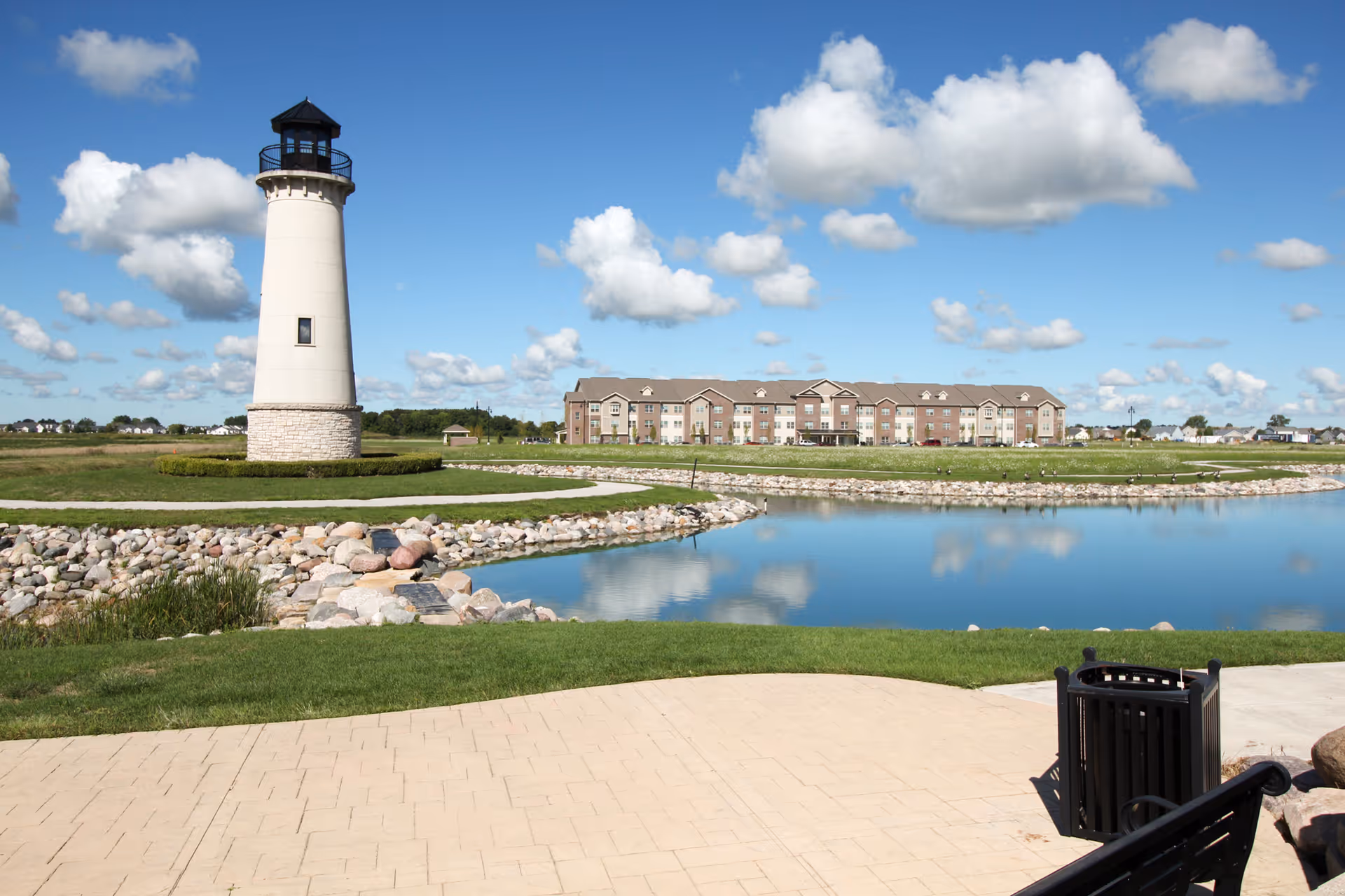 A lighthouse beside a pond with the Harbor Town Senior Residence building reflected in the water under a bright blue sky.