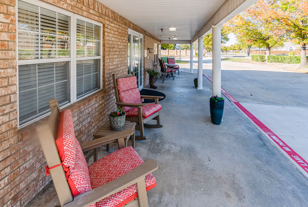 Covered outdoor patio area with several cushioned chairs and small tables along a brick wall. There are potted plants on the tables and on the ground near white columns. The patio overlooks a parking area with trees and bushes in the background.
