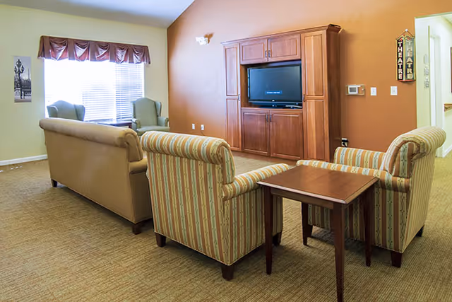 A cozy living room area with a beige sofa, two striped armchairs, a wooden side table, and two green armchairs near a window with blinds and a valance. A wooden entertainment center with a flat-screen TV is mounted on a warm-toned wall. There are signs on the wall indicating a theatre room nearby.
