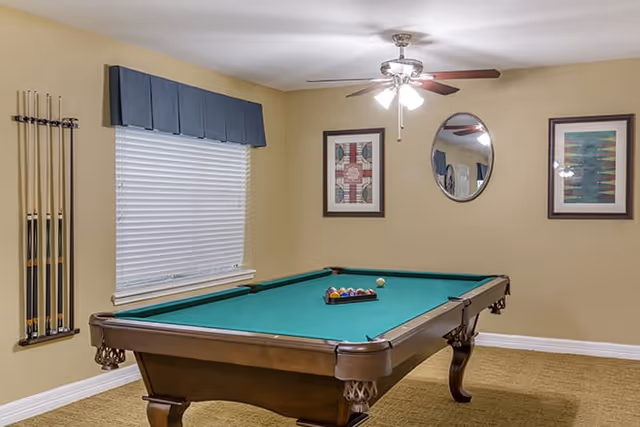 A game room with a green felt pool table set up for a game, a ceiling fan with lights above, a window with a blue valance, and three framed wall decorations including a round mirror.