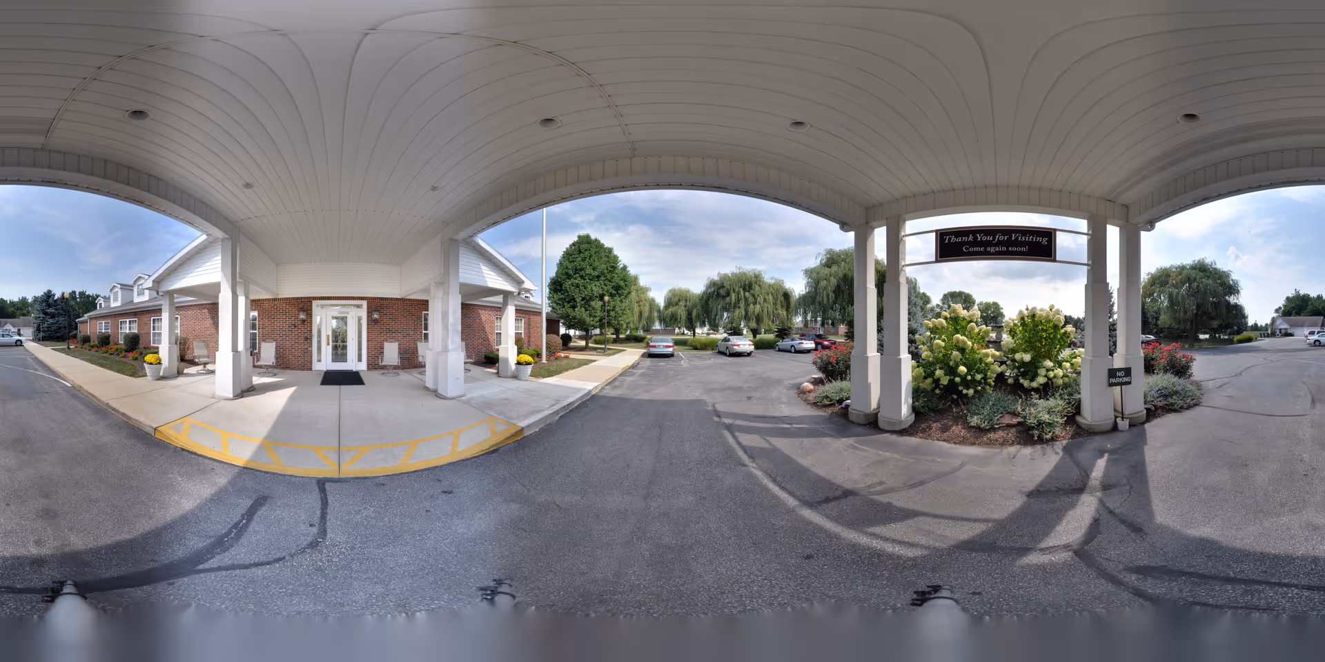 Covered entrance area of a senior living facility with a brick building on the left and a parking lot with cars and landscaped greenery on the right. There is a sign hanging from the ceiling that says 'Thank You for Visiting Come again soon.'