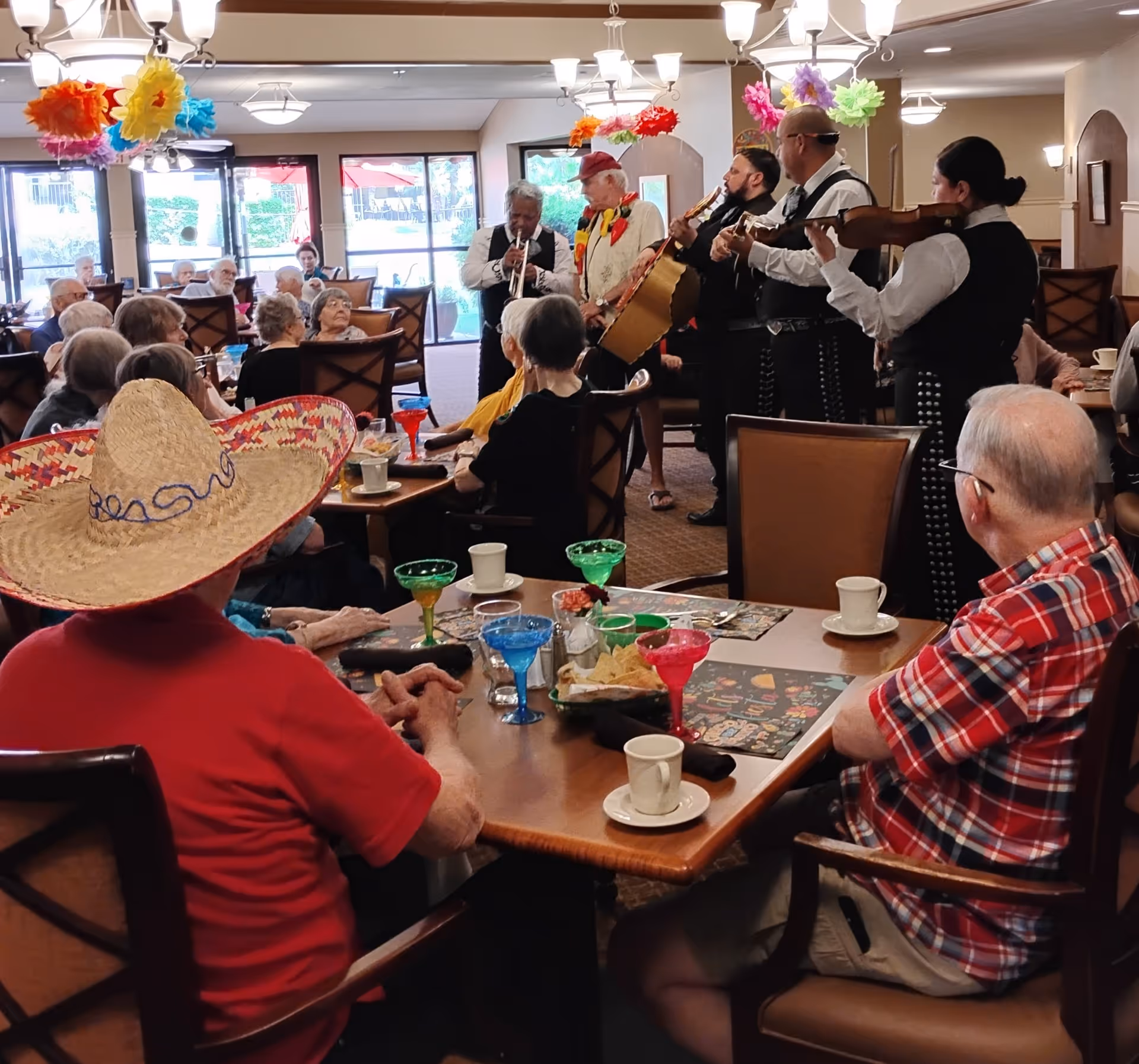 A group of elderly people seated at tables in a decorated dining room watching a mariachi band perform. The room has colorful paper flowers hanging from the ceiling and large windows in the background.