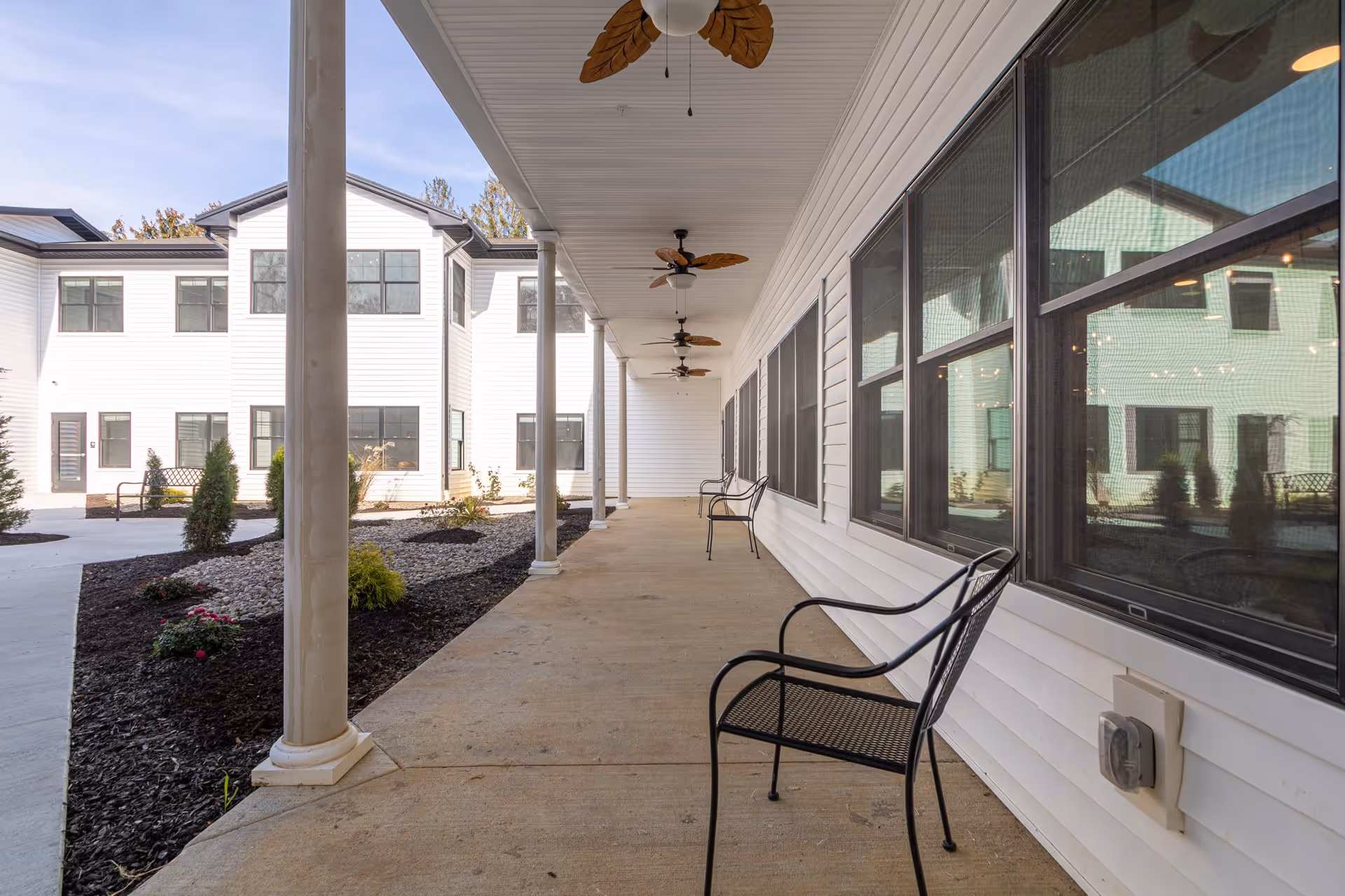 Covered outdoor walkway with ceiling fans and black metal chairs along the side of a white building. The walkway overlooks a landscaped area with small bushes and rocks, and the building has large windows reflecting the surroundings.