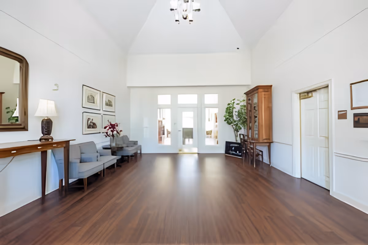 Spacious high-ceiling common room with wooden floors, seating along the left wall, a cabinet and plants on the right, and double doors at the far end.