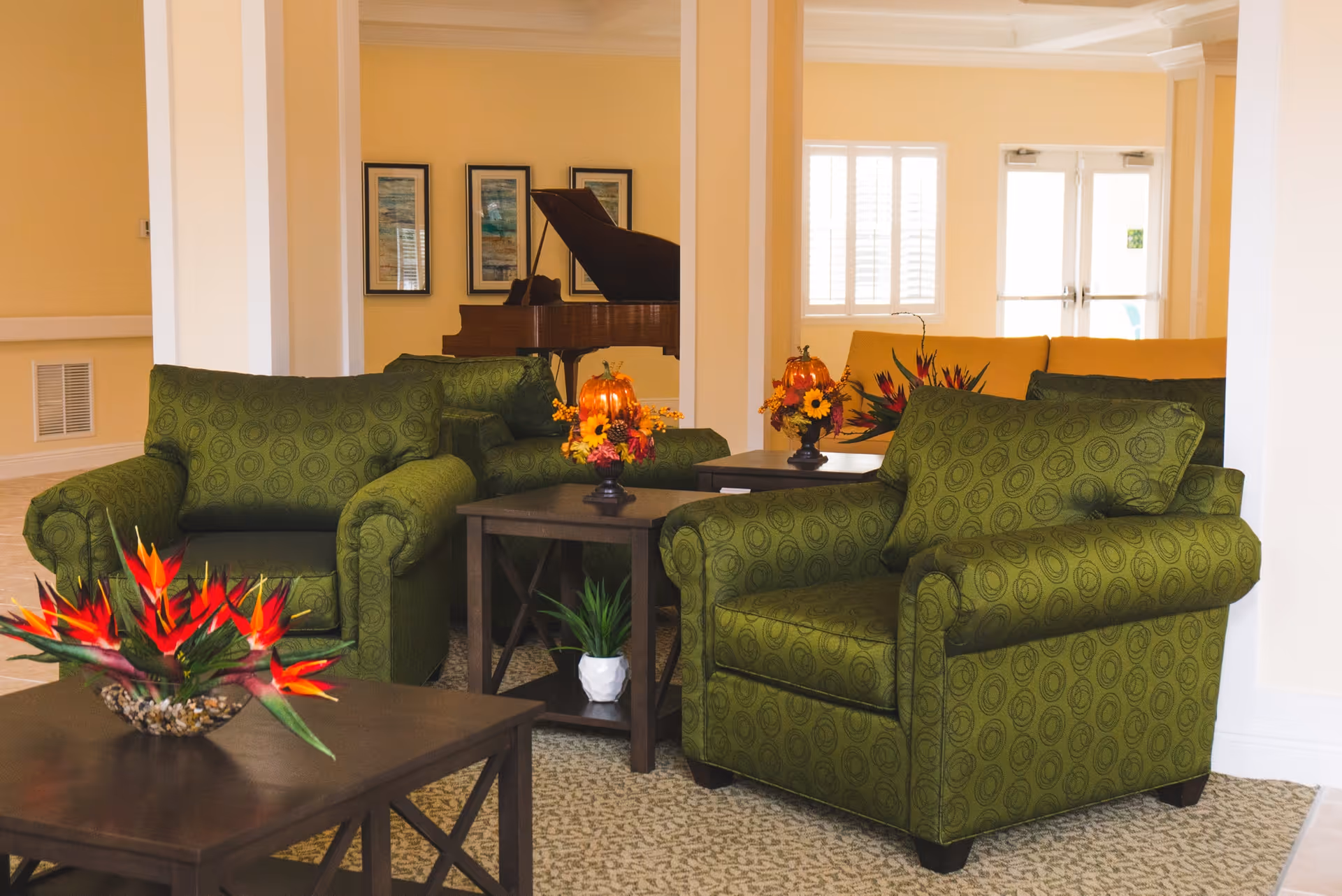 A cozy senior living common area with two green patterned armchairs, a wooden coffee table with a vibrant flower arrangement, and a grand piano in the background. The room is warmly lit with beige walls and carpeted flooring, featuring additional seating and decorative floral centerpieces on side tables.