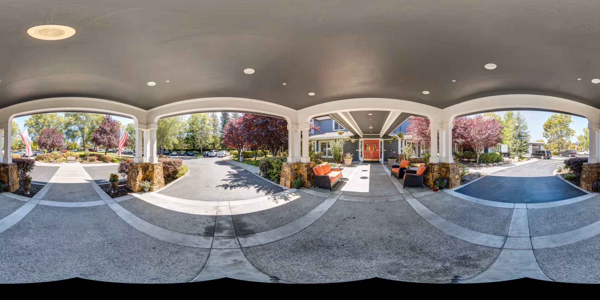 Covered entrance area of Aegis Living Moraga with seating arrangements including orange cushioned chairs and sofas. The entrance is supported by white pillars with stone bases. Surrounding the entrance are landscaped gardens with trees and shrubs, and an American flag is visible on the left side. The driveway leads to the parking area with several cars parked and more greenery in the background under a clear sky.