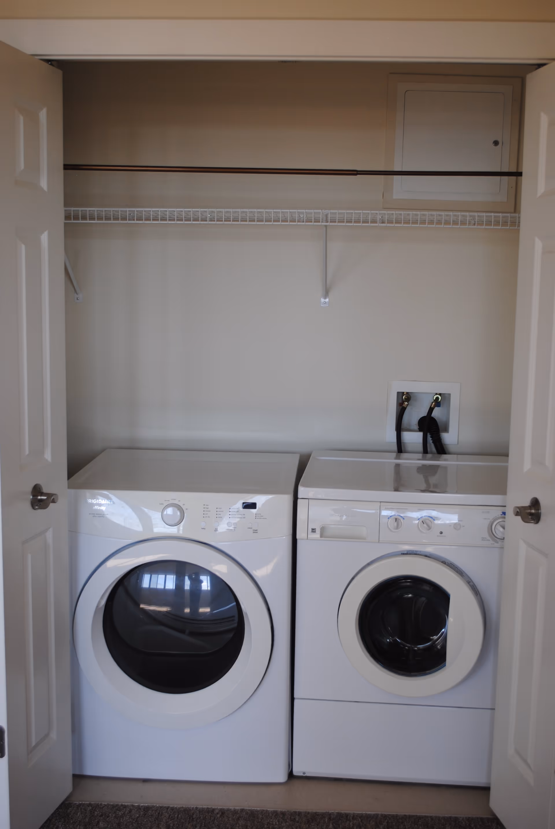 A laundry area with a white front-loading washing machine and a white front-loading dryer side by side inside a closet with double doors. Above the machines is a wire shelf and a small utility panel on the wall.
