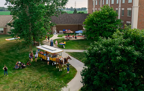 Outdoor scene at Browning Masonic Community showing a horse-drawn carriage with people gathered around it on a grassy area. In the background, there are picnic tables with umbrellas and people sitting and standing. Trees and a multi-story building are also visible.