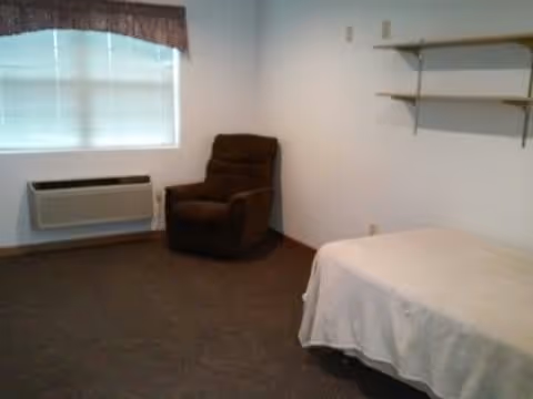 A simple bedroom with a single bed covered in a white blanket, a brown recliner chair near a window with closed blinds and a valance, a wall-mounted air conditioning unit below the window, and two wooden shelves mounted on the wall.