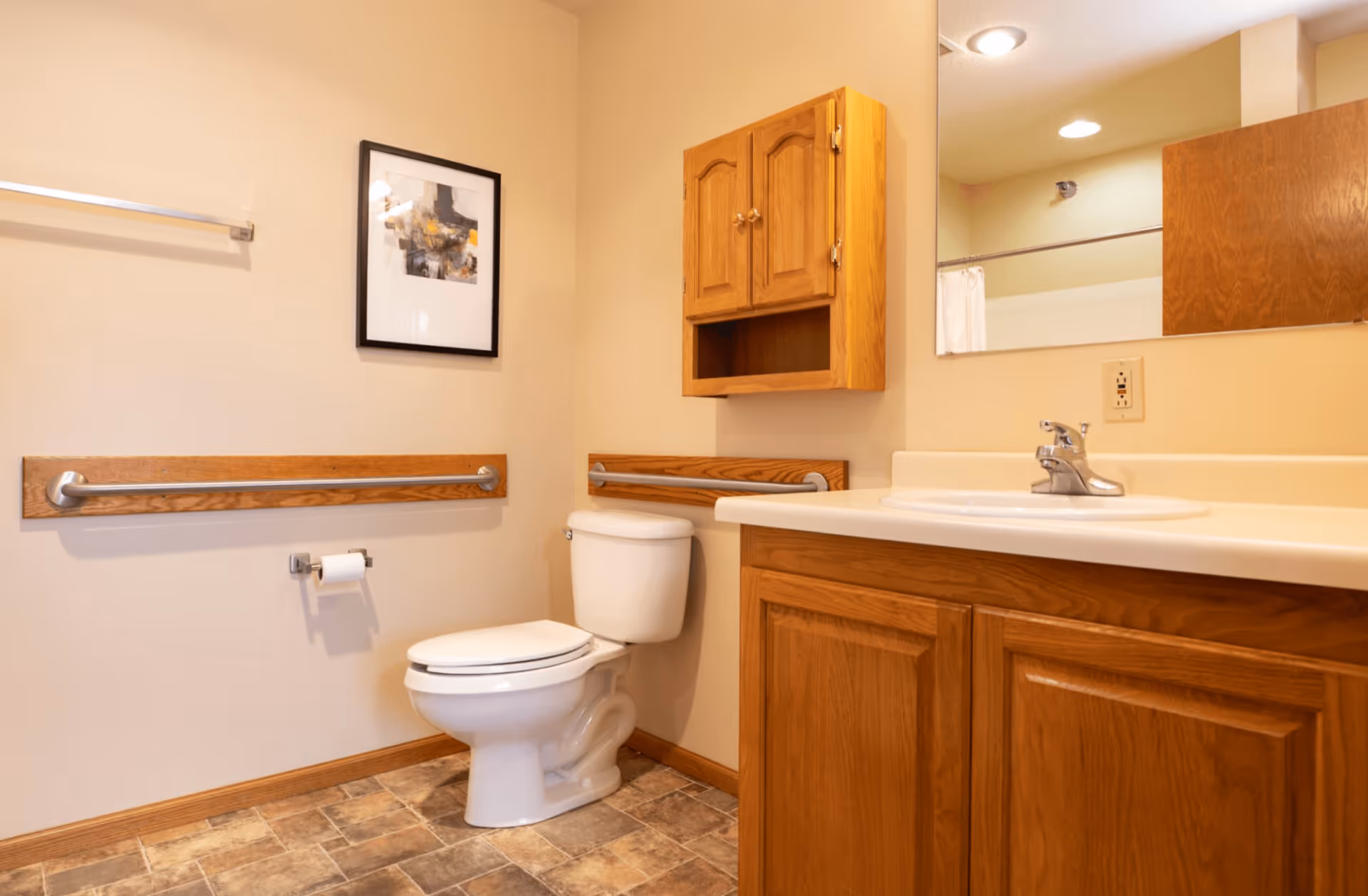 A clean bathroom with beige walls and tiled floor. It features a white toilet with wooden and metal grab bars on the walls beside it. Above the toilet is a wooden cabinet with two doors and an open shelf. To the right is a white sink with a wooden vanity cabinet below and a large mirror above. A framed abstract artwork hangs on the wall to the left.
