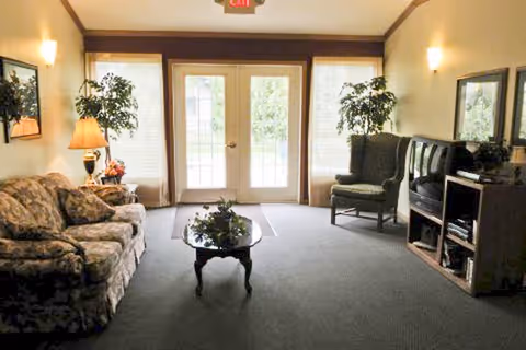 A furnished common sitting room with a floral sofa, armchair, coffee table, TV stand and double glass doors with plants on either side.