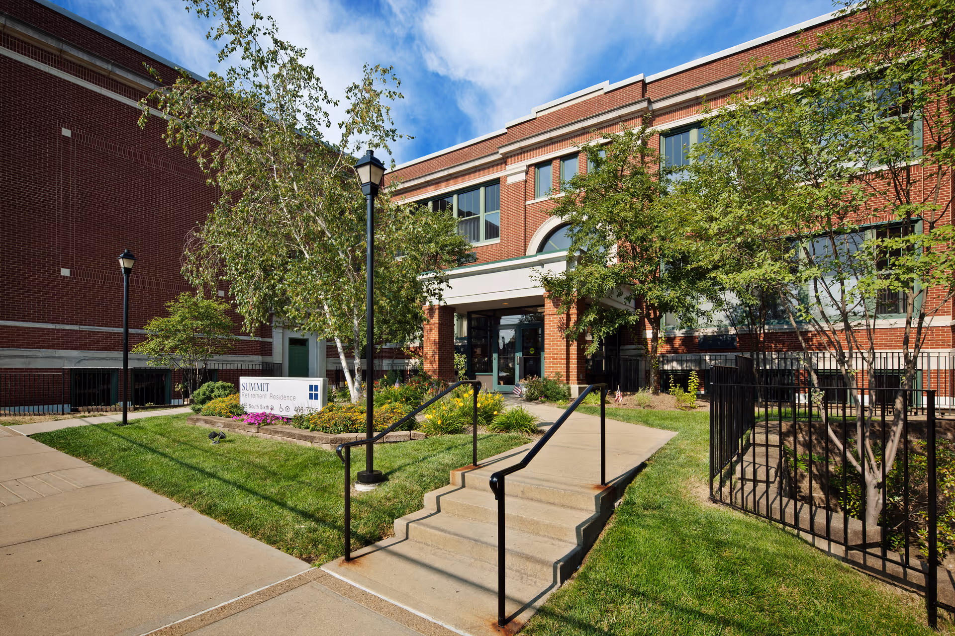 Exterior view of Summit Retirement Residence showing a brick building with large windows, a main entrance with a covered walkway, a small set of stairs with handrails, green grass, trees, and a sidewalk leading to the entrance under a partly cloudy sky.