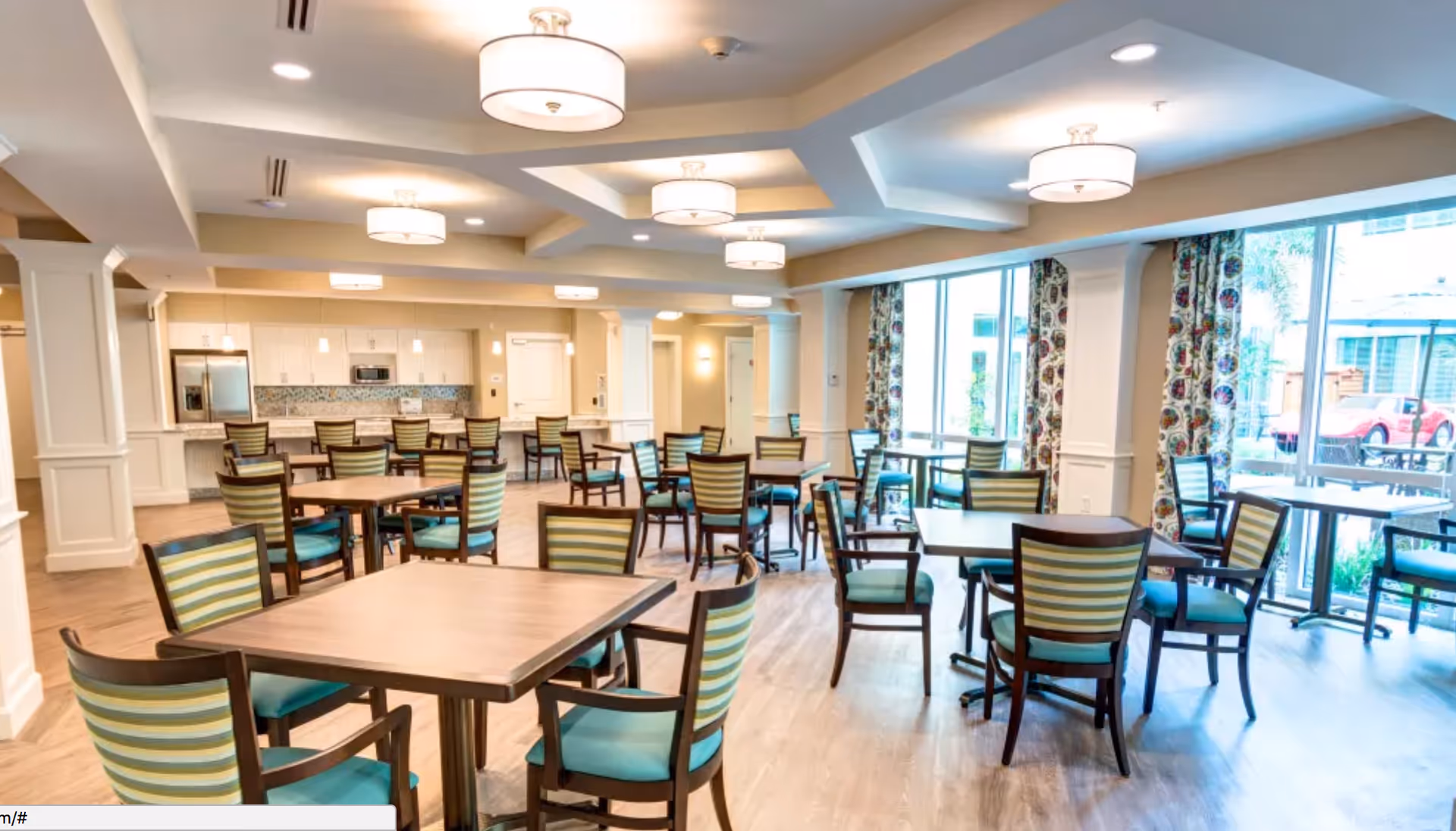 Bright dining room with multiple tables and striped chairs, large windows, and a kitchenette in the back.