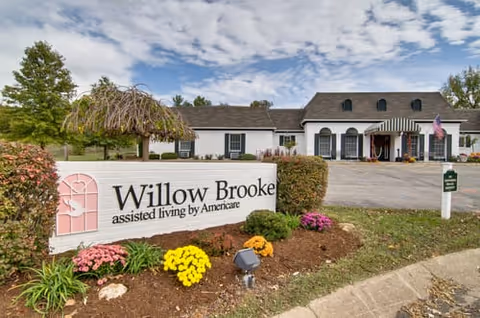 Entrance of the Willow Brooke assisted living facility with a large sign, landscaped flowers, and the building facade in the background.