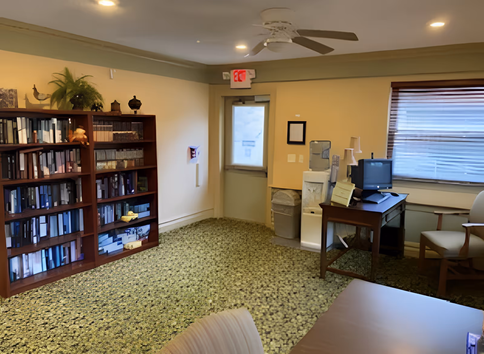 A cozy common room with bookshelves, a desk holding a small TV, seating, and a window.