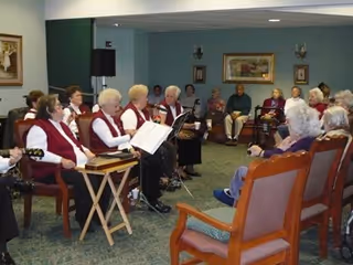 A group of elderly people seated in a semi-circle in a common room, some wearing matching red vests and holding musical instruments or songbooks, while others sit and listen attentively.