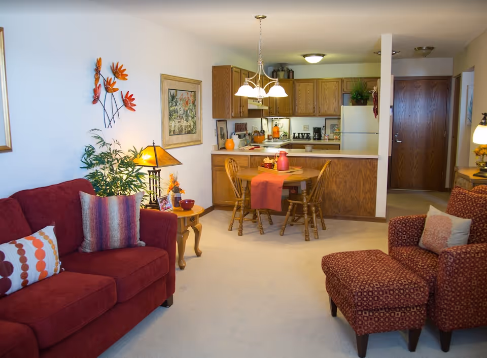 Open-plan living room with a red sofa and patterned armchair, a small wooden dining table, and a kitchen in the background.