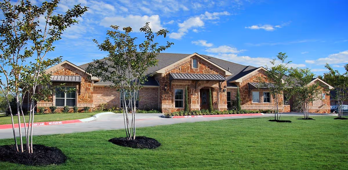 Exterior view of a single-story building with a stone and brick facade, surrounded by a well-maintained green lawn and young trees under a blue sky with scattered clouds.