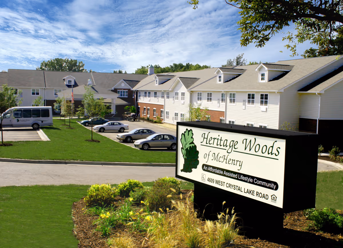 Exterior view of Heritage Woods of McHenry assisted living community showing a large two-story building with white siding and brick accents, a parking lot with several cars and a van, green lawns, trees, and a sign in the foreground with the community name and address.