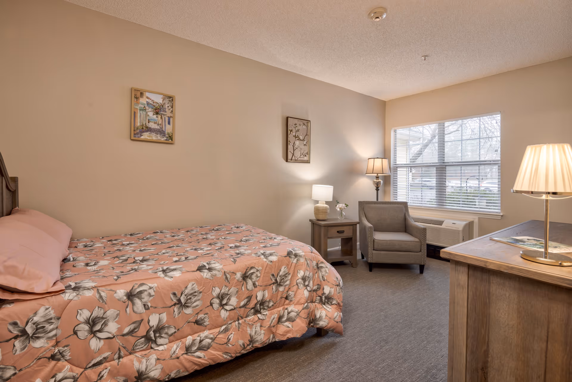 A cozy bedroom with a bed covered in a pink floral comforter, a wooden nightstand with a lamp and a small flower vase, a gray armchair near a large window with blinds, and a wooden dresser with a lamp on top. Two framed pictures hang on the beige walls.